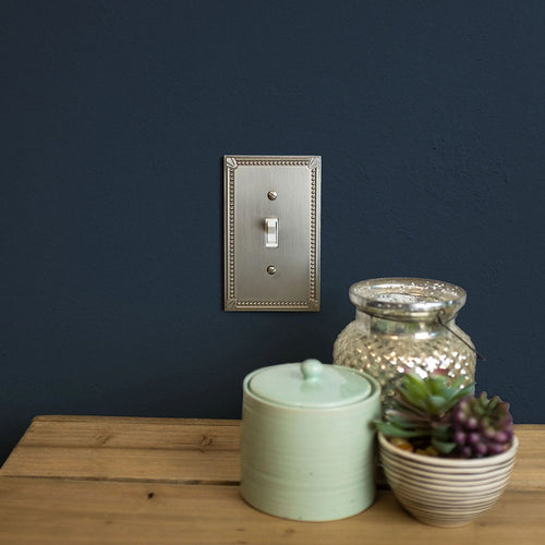 A photograph of the Imperial Bead brushed nickel wallplate on a navy wall, above a wood table, next to a green ceramic pot, and a potted cactus plant