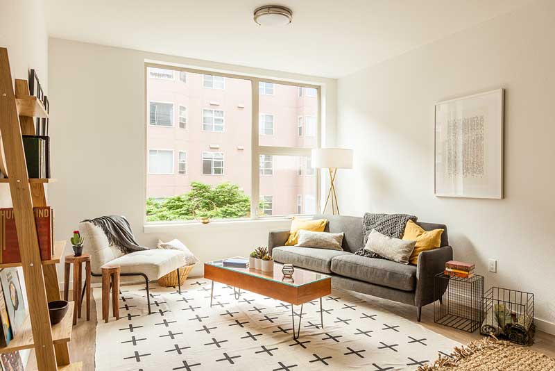 a clean apartment with white walls, a white decorative area rug, a large window letting in large amounts of natural light, and Scandinavian style couches and coffee table