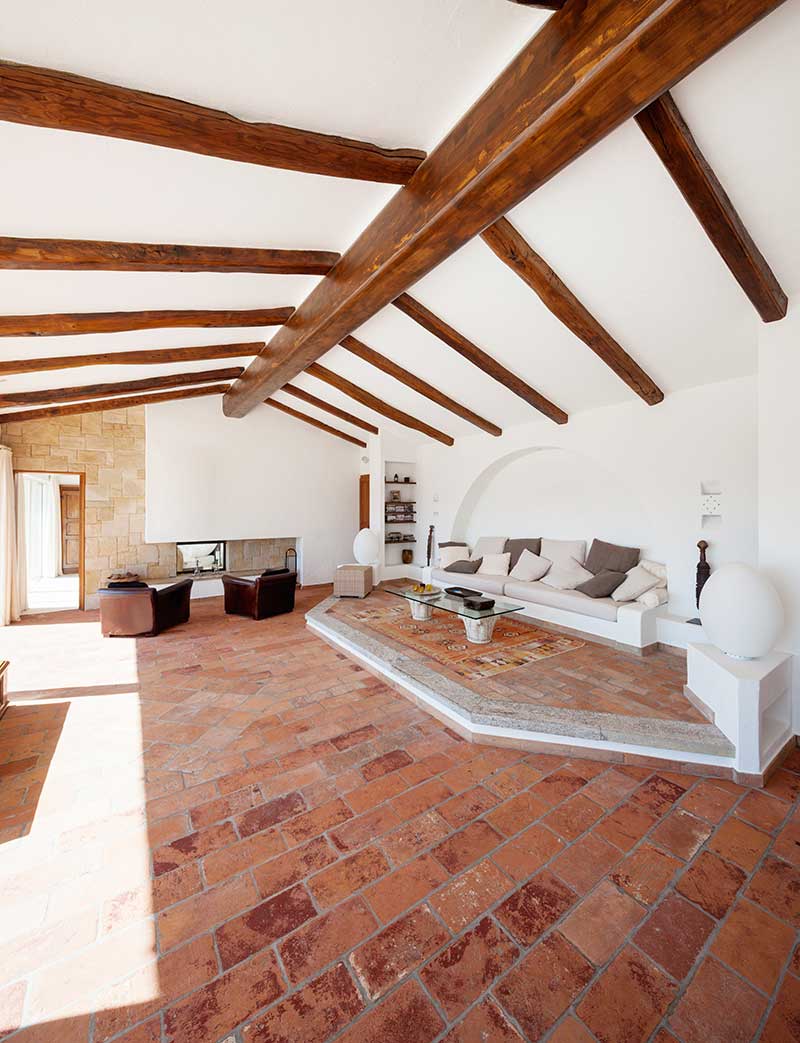 A large Southwestern decor style living room with white walls, exposed wood ceiling beams, tile stone floors, dark brown leather chairs, and a white couch