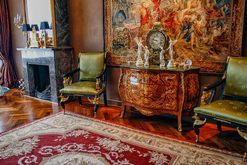 An interior photo of a professionally designed Classical decor style living room with wood floors, a black marble fireplace, a large painting on the wall, a red rug, and ornate wood furniture
