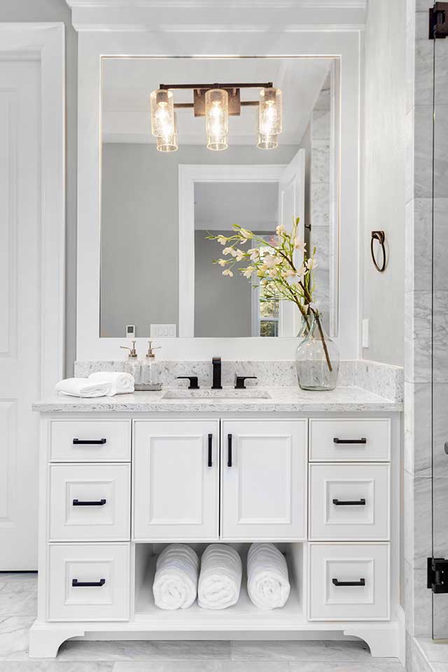 A Traditional decor style bathroom sink with a predominantly white color palette, shaker cabinets, tile floors, granite countertops, and black cabinet and sink hardware
