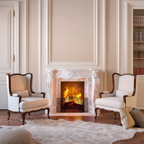 Professionally designed living room with a Classical decor style, including a large burning marble fireplace as the centerpiece of the room, two wood and fabric chairs, a bookshelf, wood floors, a fur rug, and a neutral color palette