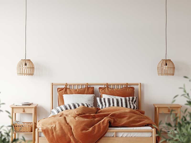 A boho bedroom with white walls, green plants, light wood furniture, woven dropdown ceiling light covers, and vibrant orange bedding