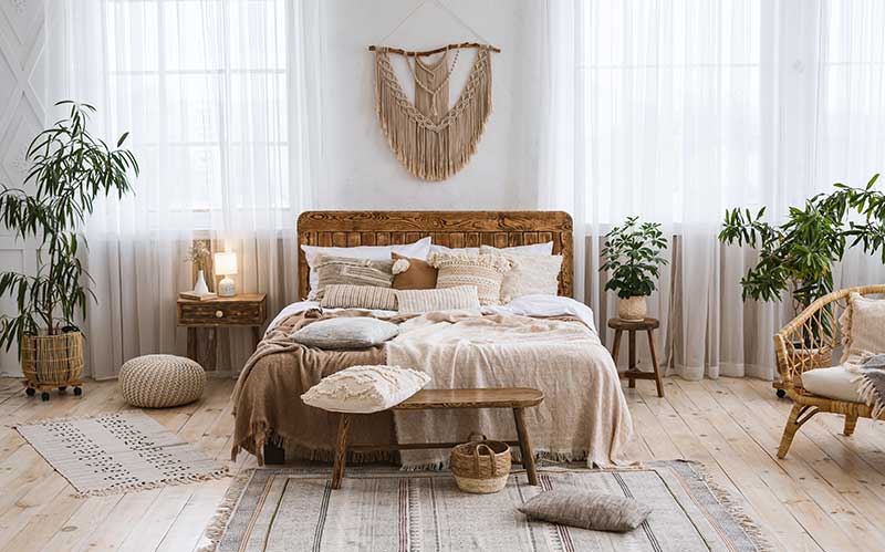 A boho bedroom with a rustic feel, raw wood plank floors, white linen window drapes, green plants, wood furniture, and a macrame wall decoration on the wall above the bed