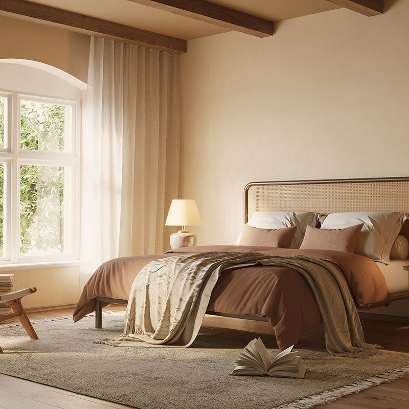 A professionally designed California Casual style bedroom with arched windows, beige walls, exposed wood ceiling beams, warm colored bedding, and a brown area rug