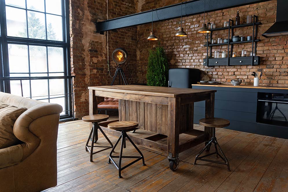 Professionally designed room with an Industrial decor style, heavy wood table, brick walls, wood floors, exposed ceiling beams, a large window, metal drop lights, and black shelving