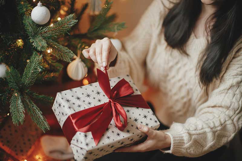 A woman in a sweater opening a Christmas present with a big red bow next to a Christmas tree