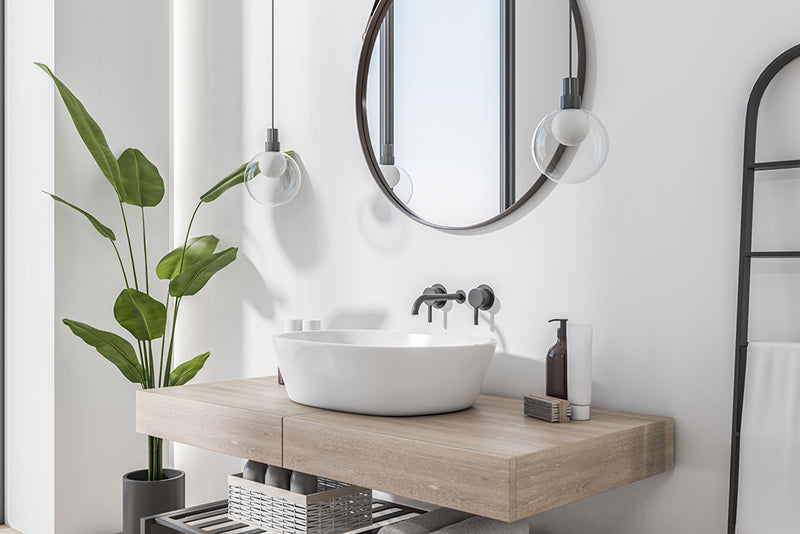 A Scandinavian decor style bathroom with white walls, black metal hardware, dropdown lights, green plants, natural light, and a light wood countertop