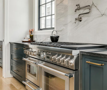 An interior photo of a professionally designed Transitional decor style kitchen with a marble backsplash, blue cabinetry, brass cabinetry hardware, a window, and a stainless steel dual oven