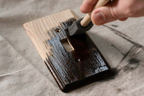 A photo of a hand holding a wood staining brush, as it lays a dark stain finish onto an unfinished Contemporary wood wallplate