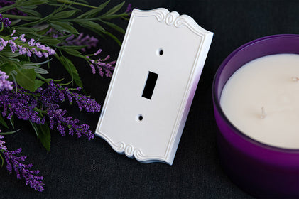 The white version of the Charleston collection of Amerelle decorative plastic wallplates on a black cloth and by a purple candle and purple lavender flowers