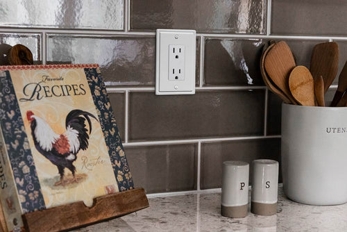 A photograph of the Moderne white metal wallplate on a tile kitchen backdrop, next to a recipe book, and other kitchen decorations