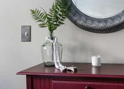 A photo of the Egg & Dart antique nickel wallplate installed on a wall, next to an antique nickel mirror, a potted fern plant, and a red table