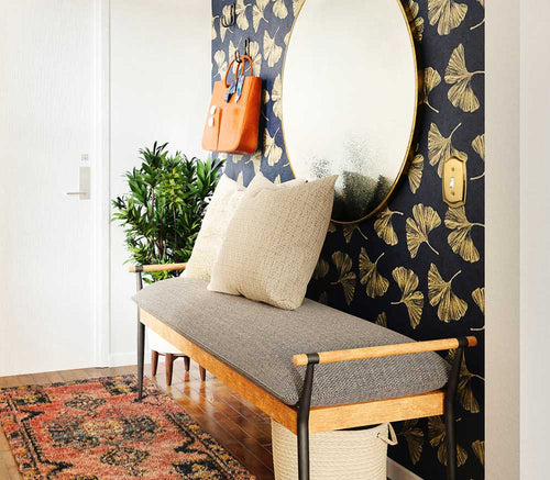 A photograph of the Sonoma polished brass metal wallplate on an entryway wall with decorative wallpaper, a round mirror, a bench, and a green potted plant