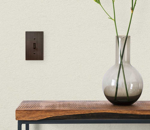 A photograph of the Elan aged bronze metal wallplate on a tan wall, next to a modern wood table, and a plant inside of a glass vase