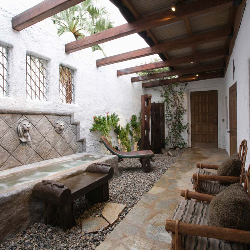 An exterior photo of a professionally designed Spanish Colonial decor style walkway and patio with large amounts of natural light, wood chairs, and a water fountain