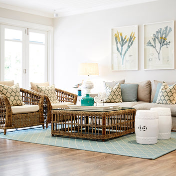 An interior photo of a professionally designed Coastal decor style living room with wicker furniture, large amounts of natural light, a blue rug, and colorful pillows