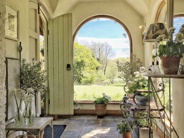 An interior photo of a professionally designed Cottage decor style entryway with a large window, wood doors, white walls, and plants on rustic tables and shelving