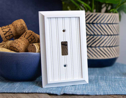 A Coastal themed photo of the Cottage White decorative wood wallplate on a wood table and a textured blue cloth by a potted plant and a bowl of wine corks