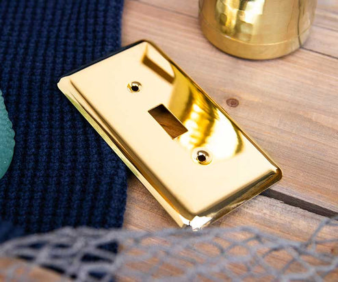 A photo of the Devon polished brass decorative metal wallplate on a wood table and a blue knitted sweater