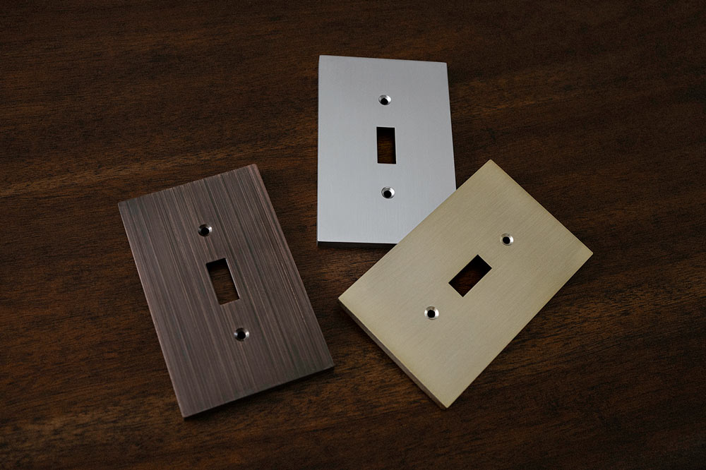 A photo of the Elan collection of metal wallplates in the brushed bronze, brushed nickel, and aged bronze color options laying down on a wood table