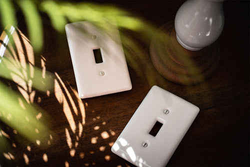 A photograph of the Metro ceramic wallplate in the biscuit and white finish options laying down on a wood table and next to a ceramic candle holder and palm leaves