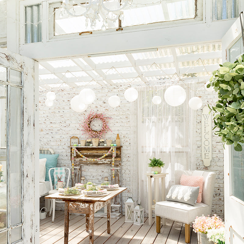 An interior photo of a professionally designed Shabby Chic French Country decor style living room with a a white color palette, white painted brick walls, handmade wood furniture, flowers, and a very large amount of natural light