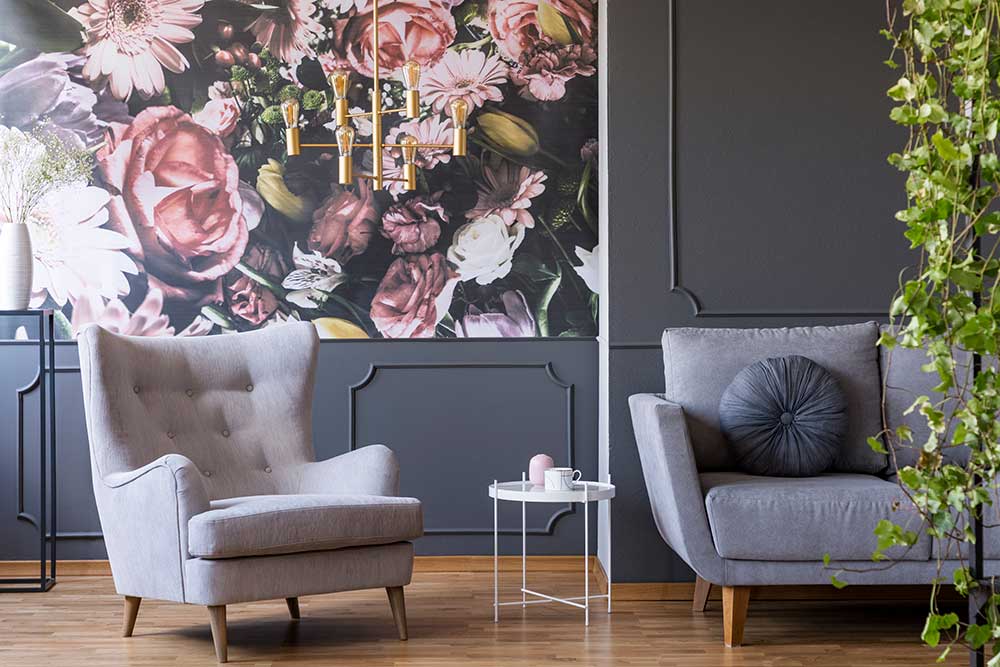 A New Traditional style room with dark grey walls, gray cloth furniture, a brass dropdown chandelier, and a wall with a detailed floral wallpaper