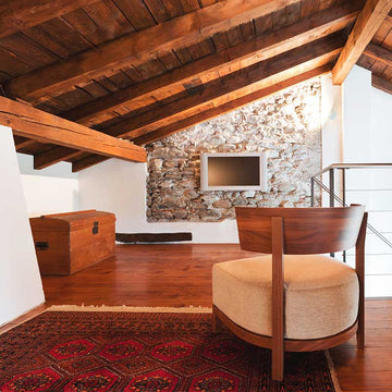 An interior photo of a professionally designed Southwestern decor style loft with a lounge chair, a flat screen tv, and a wood storage chest, wood floors, an exposed wood ceiling, and both white and stone walls