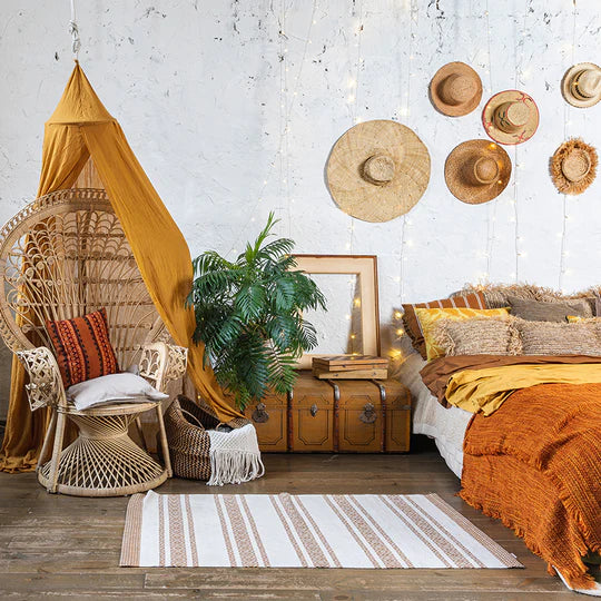 An interior photo of a professionally designed Bohemian decor style bedroom with a wicker chair, a knitted rug, a leather chest, sun hats hanging on the wall, and a bed with orange and yellow bedding