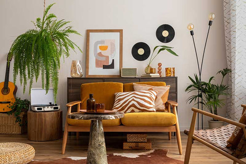 A room of various decor styles, with wood floors, white walls, green plants, a mid-century modern style lounging chair, contemporary artwork, and patterned decorative pillows