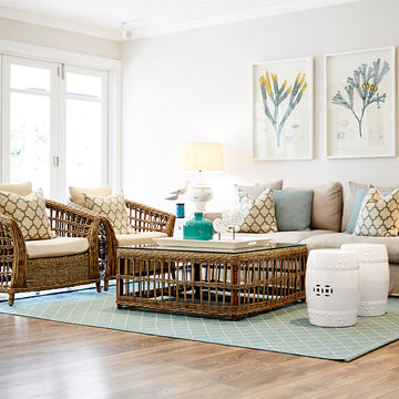 An exterior photo of a professionally designed Coastal decor style living room with wicker chairs, couch, and coffee table, a light blue rug, large windows, and colorful pillows