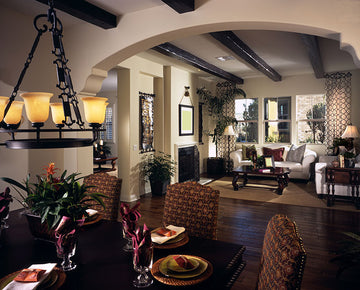 A dark, professionally designed living room and dining room with a Spanish Colonial decor style with exposed wood beams, a black iron chandelier, wood floors, and small windows