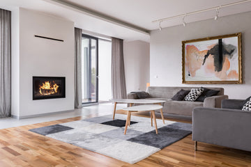 An interior photo of a professionally designed Contemporary decor style living room with a fireplace, grey couches, wood floors, a geometric style rug, and large windows with natural light