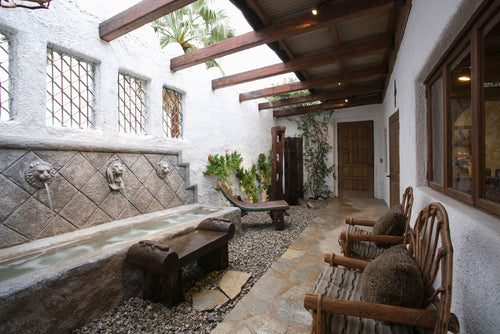An exterior photo of a professionally designed Spanish Colonial decor style walkway and patio with large amounts of natural light, wood chairs, and a water fountain