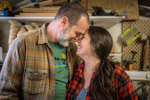 Erin and Gordon, artists and woodworkers, smiling with their arms around each other and their foreheads pressed together in the wood shop.