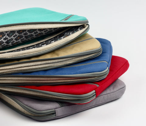A pile of brightly coloured laptop cases sits on a white background. The top one (teal outer) is partly open, showing the bold geometric Dhaka fabric lining of the case.