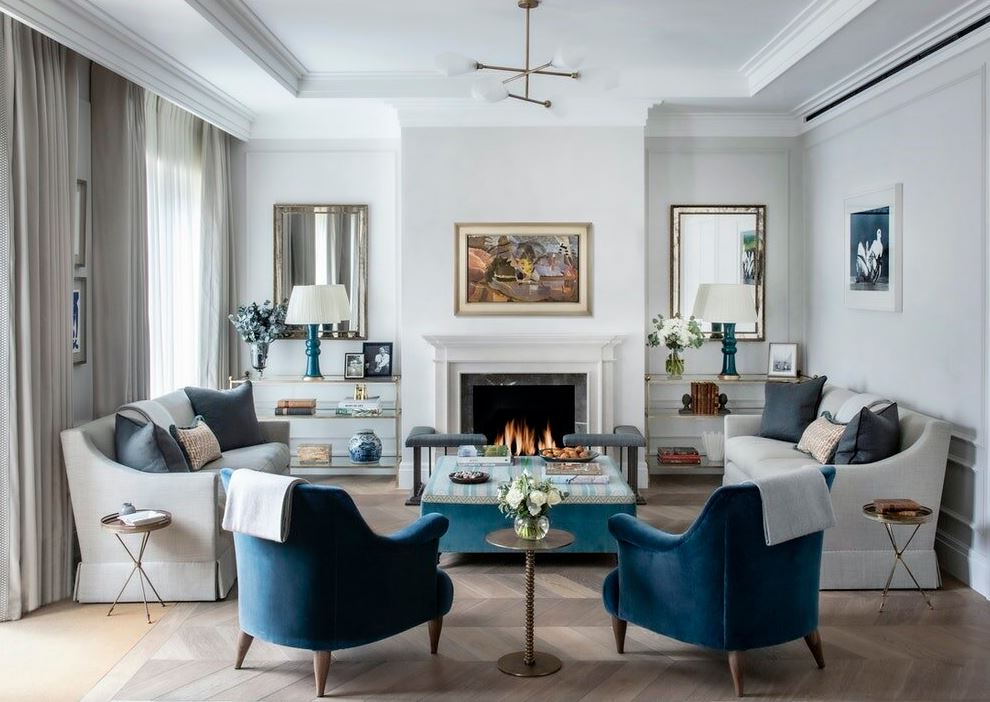 Classic living room with blue lounge chair, grey sofa, classic fireplace, modern chandelier, glass shelves with home decors, ceiling moldings and wood plank floor