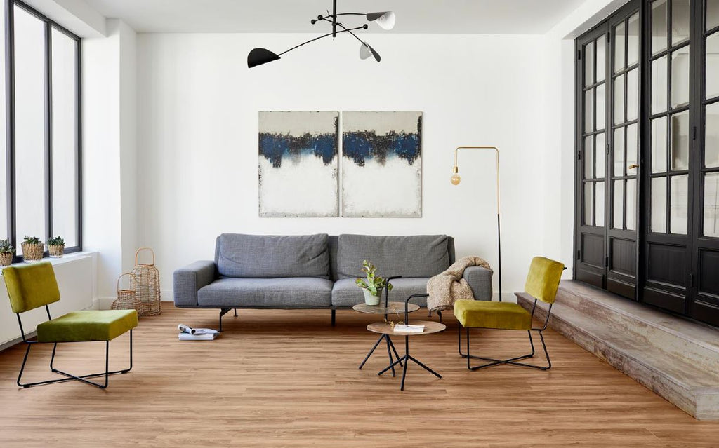 Living are with wooden plank floor, grey couch, yellow accent chairs, abstract blue and white painting, gold floor lamp, white painted wall and French folding doors