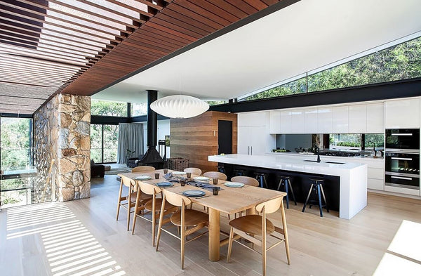Kitchen and dining room with wood dining table and chairs, glossy kitchen cabinets, white marble island countertop, wood floor planks, wood ceiling slats, cobble stones accent and large glass windows