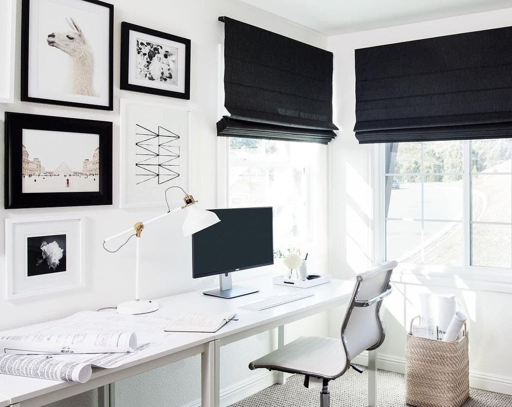 Black and white home office concept with white office desk and chair, black fabric window roman blinds, picture frames, computer monitor, weaved basket and brown carpet floor