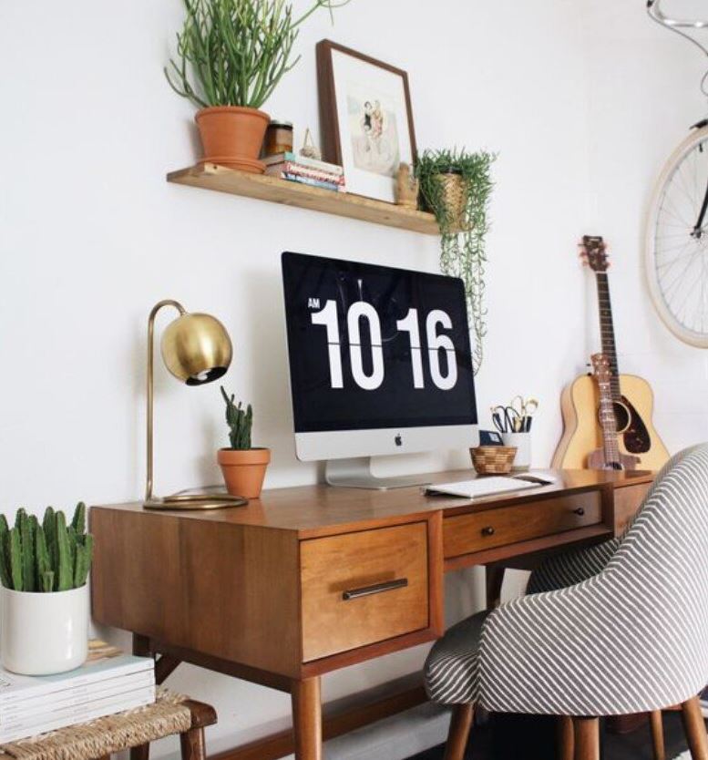 Home office showing a wood table, wall shelf, gold table lamp, upholstered seat, guitar, books, iMac computer monitor, plants and white area rug