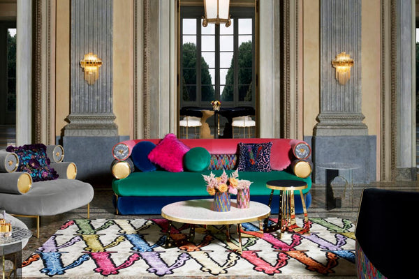 Versace's Home Collection of 2020 showing clue green and pink color combination for sofa grey lounge chairs decorative pillows on colorful printed and gold side tables and coffee tables