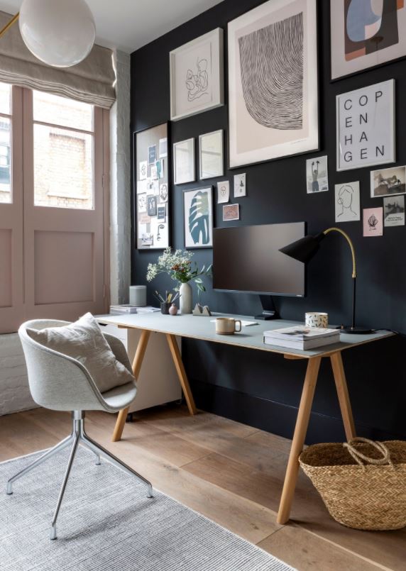 Home office with black painted wall, wood office desk, upholstered office chair, monitor, home decors, pink door, wood plank floor and rattan basket