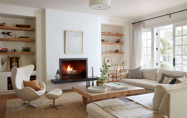 Living area with white fireplace upholstered sofa and lounge chairs neutral color are rugs decorative pillows wood shelves and white French windows