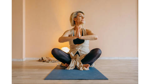 A mature woman with short blonde hair is seated in a lotus position on a yoga mat, practicing meditation.