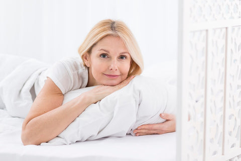 A smiling mature woman with blonde hair lies on her stomach on a white bed, resting her head on her hands atop a pillow.