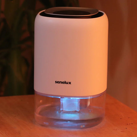 A Senelux Q4 Home Dehumidifier sitting on a table with the blue LED light showing in the clear, high capacity water tank at the bottom of the dehumidifier