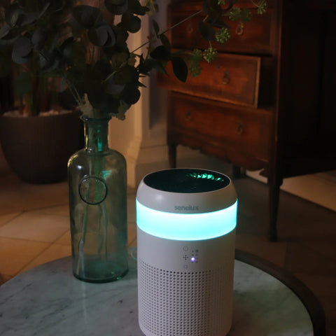 A photo of the Senelux Demi Air Purifier sitting on top of a marble effect table with a plant behind it and a pale blue LED light coming from the light strip