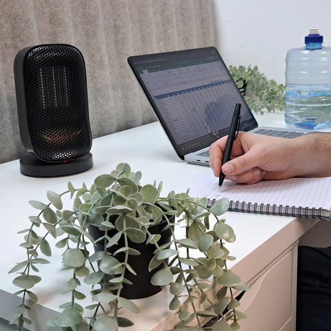A photo of a Senelux Desk Top Mini Space Heater sitting on a desk with a plant in the middle of the picture and a hand working on some paper on the desk on the right of the image